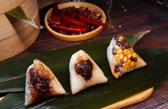 Close-up view of various Asian rice dumplings, beautifully presented on a leaf with spicy red peppers in the background.